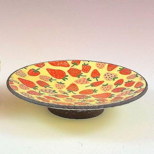 May include: A ceramic plate featuring a yellow background adorned with red strawberry illustrations. The plate has a dark grey rim and a brown base. Ideal for serving food or as a decorative kitchen item.