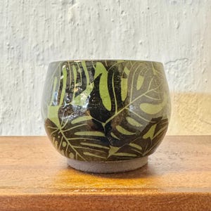 May include: A green ceramic cup with a black leaf pattern. The cup has a rounded shape and a flat base.