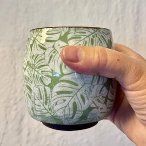 May include: A rounded ceramic cup featuring a repeating pattern of white monstera leaves against a sage green background. The cup has a smooth, curved silhouette with a dark brown rim and a dark brown base, showcasing a detailed botanical print.
