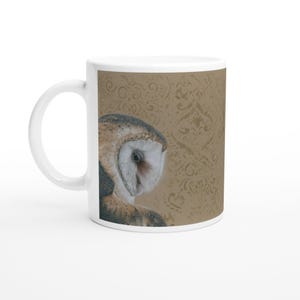 Könnte beinhalten: Ein weißer Keramikbecher mit einer detaillierten Illustration einer Schleiereule mit weißen, braunen und schwarzen Federn. Die Eule ist auf der linken Seite vor einem beigefarbenen Hintergrund mit einem dezenten Damastmuster abgebildet.