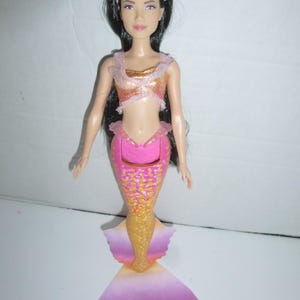 May include: A mermaid doll with long black hair, wearing a pink headband, and a two-piece outfit. The top is gold and pink, and the mermaid tail is pink, gold, and orange. The doll has a fair complexion and is posed upright.