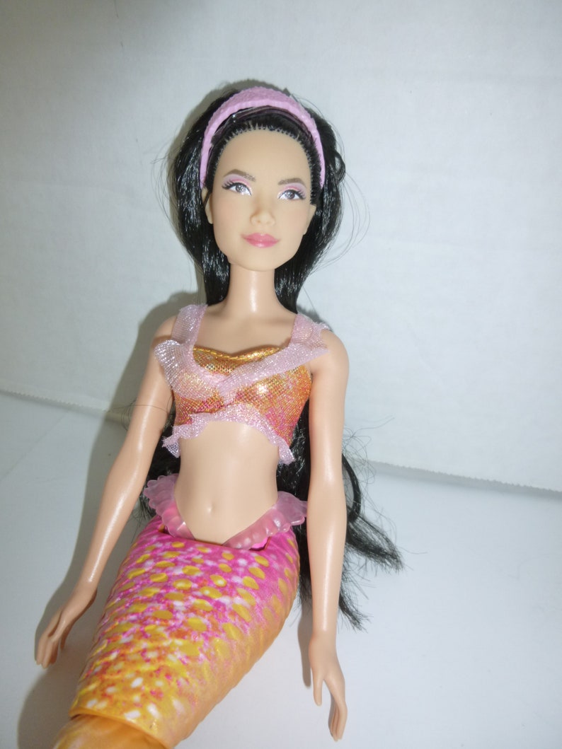 May include: A mermaid doll with long black hair and a pink headband. The doll wears a sparkly orange and pink top and a matching mermaid tail. The tail has a scale pattern in shades of yellow, orange, and pink.