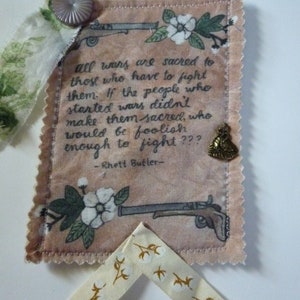 May include: A fabric wall hanging with a quote by Rhett Butler, featuring floral and gun illustrations. The quote reads, "All wars are sacred to those who have to fight them." The hanging has a decorative button and ribbon.
