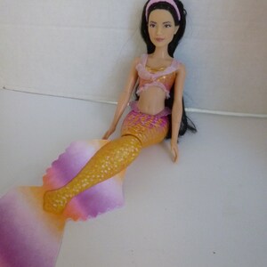 May include: A mermaid doll with long black hair, wearing a pink headband and a two-piece outfit. The top is gold and pink, and the mermaid tail transitions from gold to pink and purple.