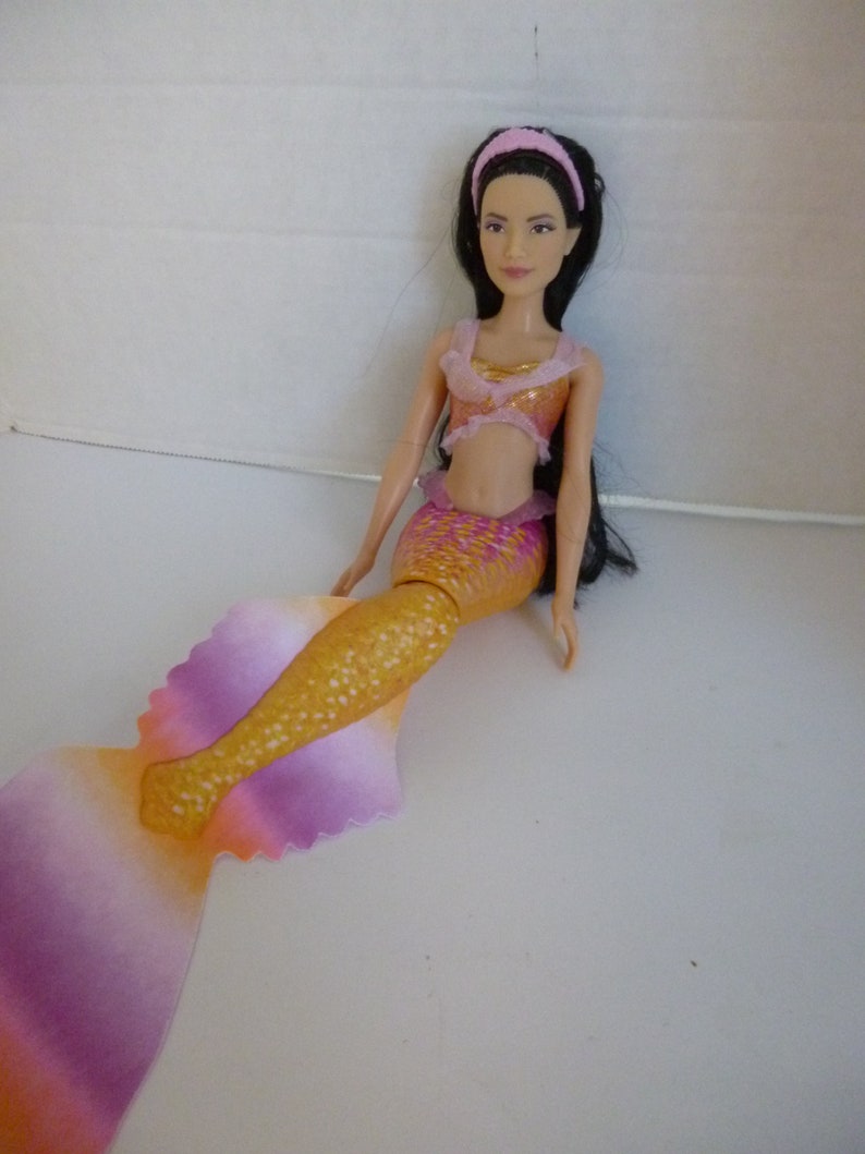 May include: A mermaid doll with long black hair, wearing a pink headband and a two-piece outfit. The top is gold and pink, and the mermaid tail transitions from gold to pink and purple.