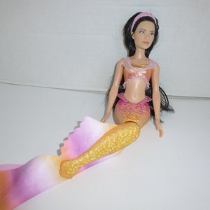 May include: A mermaid doll with long black hair, wearing a pink headband, and a sparkly gold and pink top. The doll has a pink and gold mermaid tail with a flowing, multi-colored fin. The doll is sitting on a white surface.