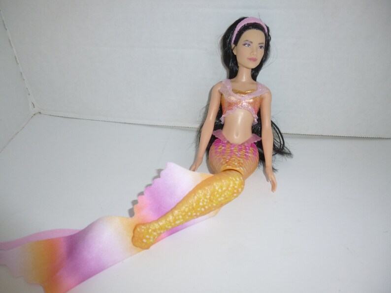 May include: A mermaid doll with long black hair, wearing a pink headband, and a sparkly gold and pink top. The doll has a pink and gold mermaid tail with a flowing, multi-colored fin. The doll is sitting on a white surface.