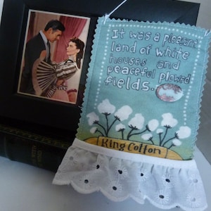 May include: A blue and white fabric bookmark with a quote about a pleasant land of white houses and peaceful plowed fields. The bookmark has a white lace trim and the text "King Cotton" on a yellow banner.
