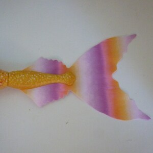 May include: A mermaid doll with a vibrant, colorful design. The tail features a gradient of orange, purple, and pink, with a yellow-gold midsection. The doll's body is pink with yellow spots, and it has black hair.