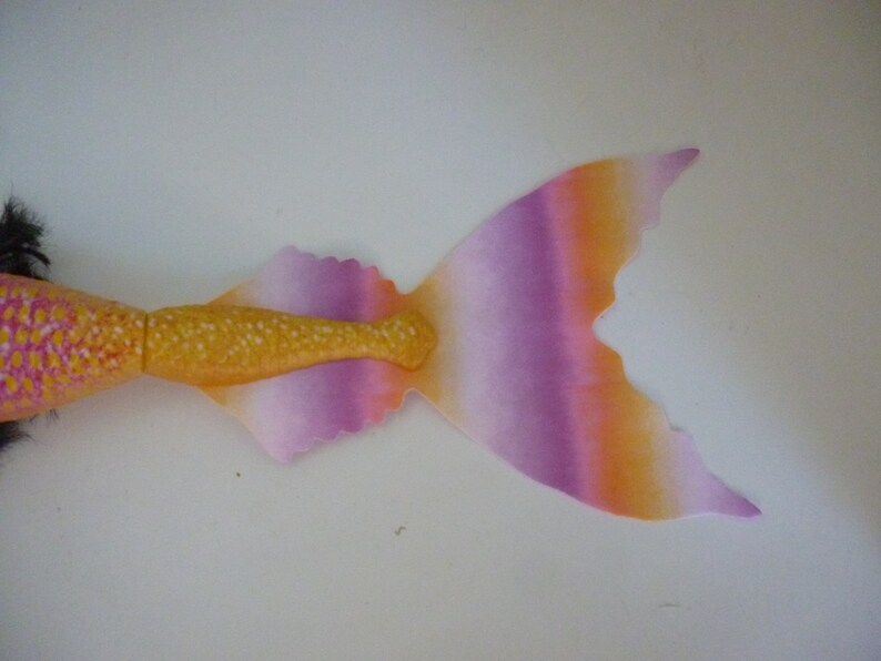 May include: A mermaid doll with a vibrant, colorful design. The tail features a gradient of orange, purple, and pink, with a yellow-gold midsection. The doll's body is pink with yellow spots, and it has black hair.