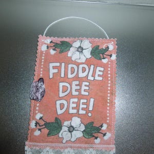 May include: A pink fabric sign with white lace trim and white flowers. The sign says "Fiddle Dee Dee!"