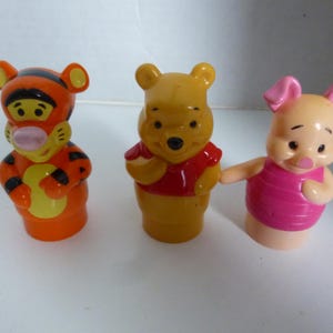May include: Three plastic figurines of Winnie the Pooh characters: Tigger, Winnie the Pooh, and Piglet. Tigger is orange with black stripes, Winnie the Pooh is yellow with a red shirt, and Piglet is pink with a pink shirt.
