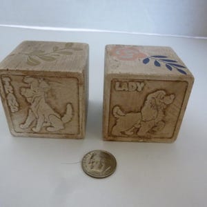May include: Two vintage wooden blocks with dog illustrations. One block reads "TRIX" and the other reads "LADY". The blocks have a light brown color with floral and leaf designs on top. A dime is placed in front of the blocks.