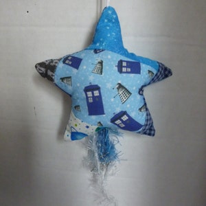 May include: A blue and white star-shaped ornament with a blue and white tassel. The star is decorated with a pattern of blue police boxes and Daleks.