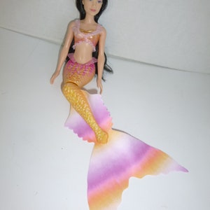 May include: A mermaid doll with long black hair and a pink headband. The doll wears a gold and pink top and a matching mermaid tail. The tail transitions from yellow to pink and orange. The doll is a toy.