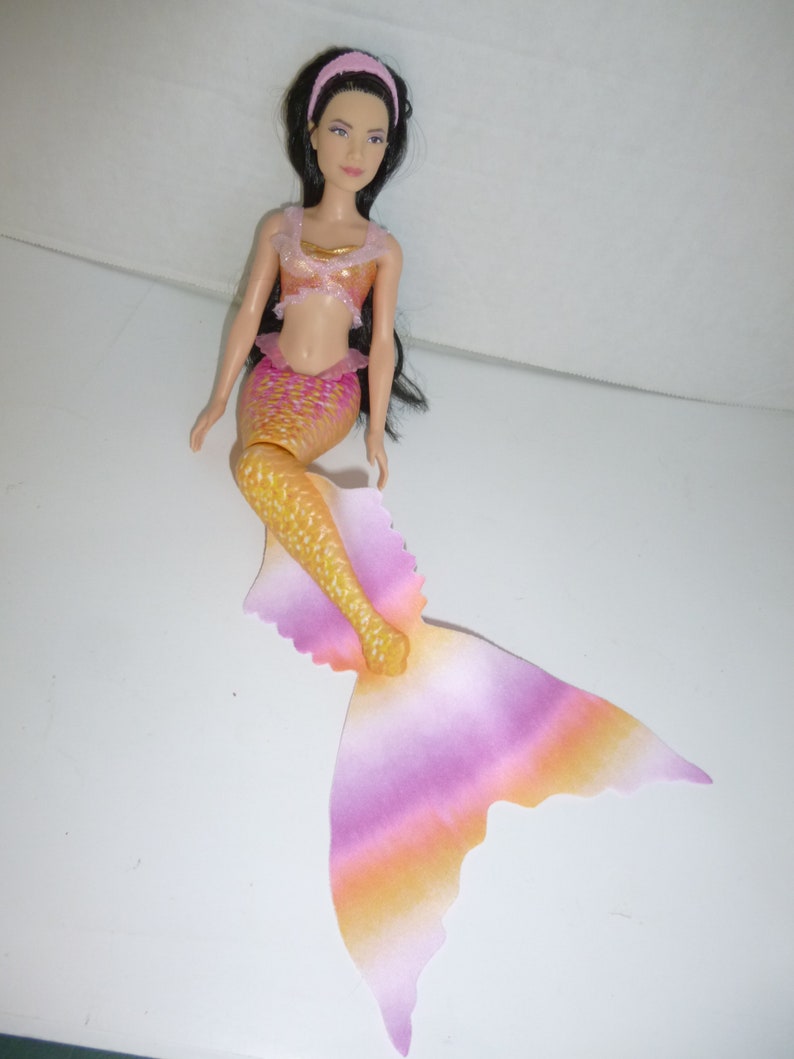 May include: A mermaid doll with long black hair and a pink headband. The doll wears a gold and pink top and a matching mermaid tail. The tail transitions from yellow to pink and orange. The doll is a toy.