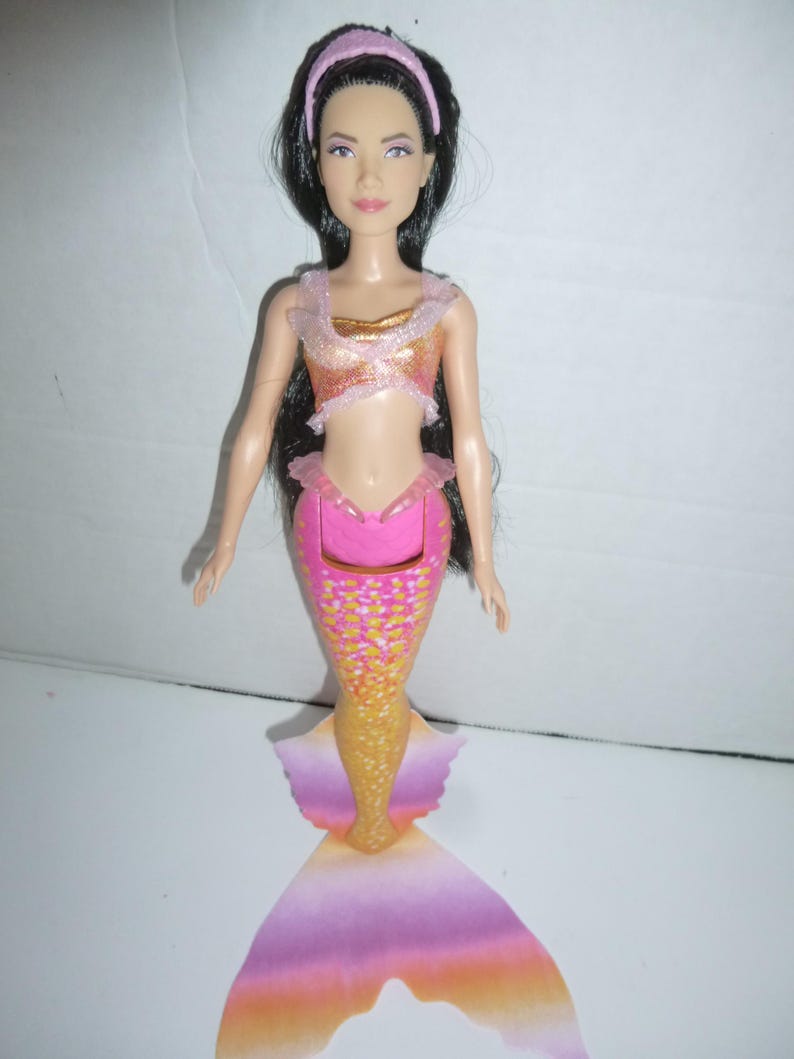 May include: A mermaid doll with long black hair, wearing a pink headband, and a two-piece outfit. The top is gold and pink, and the mermaid tail is pink, gold, and orange. The doll has a fair complexion and is posed upright.