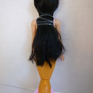 May include: A doll with long black hair, a pink top, and a tan mermaid tail with pink fins. The doll's hair is secured with clear bands. The mermaid doll is viewed from the back.