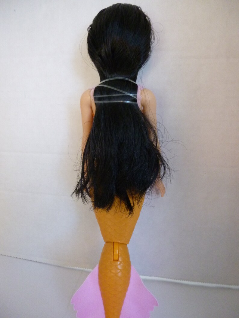 May include: A doll with long black hair, a pink top, and a tan mermaid tail with pink fins. The doll's hair is secured with clear bands. The mermaid doll is viewed from the back.