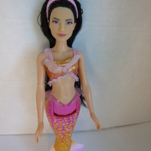 May include: A mermaid doll with long black hair and a pink headband. The doll wears a pink and gold top and a pink, orange, and yellow mermaid tail. The tail has a fin at the end. The doll is a toy.