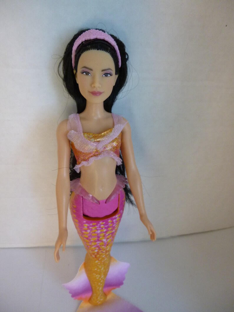 May include: A mermaid doll with long black hair and a pink headband. The doll wears a pink and gold top and a pink, orange, and yellow mermaid tail. The tail has a fin at the end. The doll is a toy.