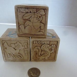 May include: Three wooden toy blocks with carved images of Disney characters. One block features Thumper the rabbit, another Bambi the deer, and the third Flower the skunk. The blocks are light brown and appear to be vintage.