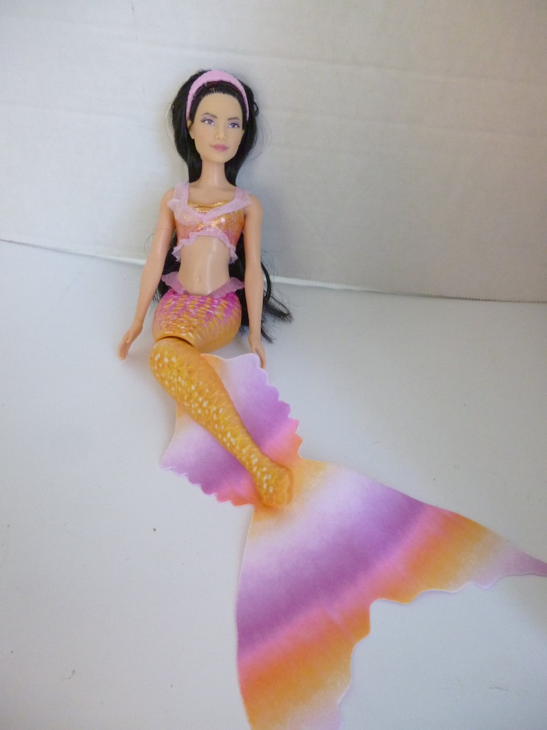 May include: A mermaid doll with long black hair, a pink headband, and a gold and pink tail. The doll is wearing a pink and gold bikini top. The tail is a gradient of pink, orange, and yellow.