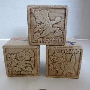 May include: Three vintage wooden blocks featuring embossed images of Pinocchio, Jiminy Cricket, and Figaro. Each block has the character's name embossed below the image. The blocks are beige and appear to be well-preserved.