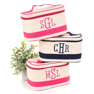 May include: Three personalized cosmetic bags stacked on top of each other. The bags are cream-colored with a colored stripe and matching zipper. Each bag has embroidered initials in a contrasting color. One bag has pink accents and initials, another has navy accents and initials, and the third has pink accents and initials.
