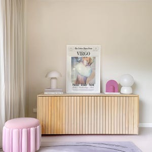 Op de afbeelding: Een licht houten kast met verticale lamellen bevat een ingelijste "Virgo" print, een witte paddenstoellamp en decoratieve objecten. Een roze, gecanneleerde poef staat op een lichtroze vloerkleed. De kamer heeft een neutraal kleurenpalet.
