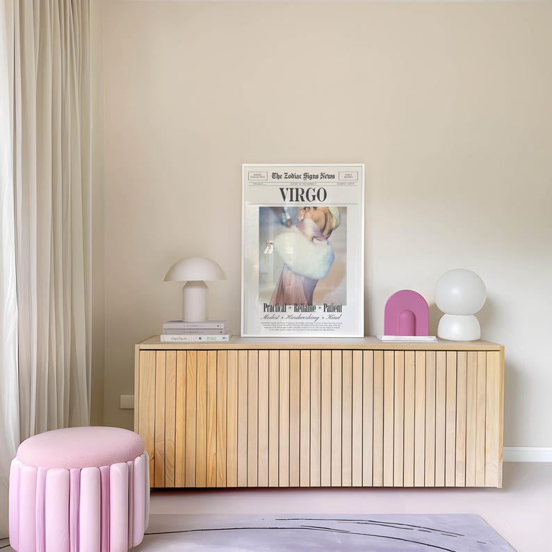 Op de afbeelding: Een licht houten kast met verticale lamellen bevat een ingelijste "Virgo" print, een witte paddenstoellamp en decoratieve objecten. Een roze, gecanneleerde poef staat op een lichtroze vloerkleed. De kamer heeft een neutraal kleurenpalet.