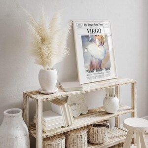 Op de afbeelding: Een rustieke houten plank toont een ingelijste "Virgo" dierenriemteken print, een witte vaas met pampasgras, boeken en decoratieve manden. De print toont een vrouw met een bontstola. Een kleine kruk en een grote witte vaas zijn ook zichtbaar.