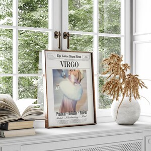 Op de afbeelding: Een ingelijste afdruk van een krantenpagina getiteld "The Zodiac Signs News" met het sterrenbeeld "Virgo". De afbeelding toont een vrouw in een roze jurk en witte bontjas. De print wordt weergegeven op een witte vensterbank met een vaas en boeken.