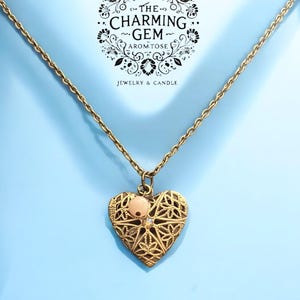 May include: A gold heart-shaped locket necklace with a delicate filigree design. The locket has a small pink stone in the centre. The necklace is on a light blue background.