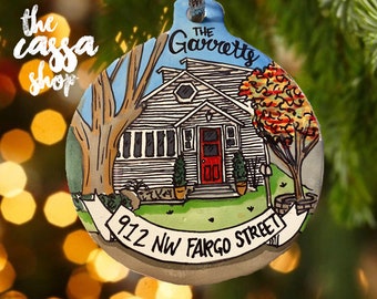 Könnte beinhalten: Ein rundes Keramikornament mit einer handgemalten Illustration eines Hauses mit weißer Fassade und roter Tür. Der Text lautet The Garretts und 912 NW FARGO STREET. Ein großer Baum und ein kleinerer Baum mit Herbstlaub sind zu sehen.
