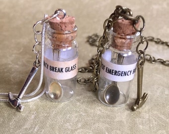 May include: Two glass bottle necklaces with cork stoppers. One has a silver chain and a hammer charm, the other a bronze chain and hammer charm. Each bottle has a label that reads "Emergency Break Glass".