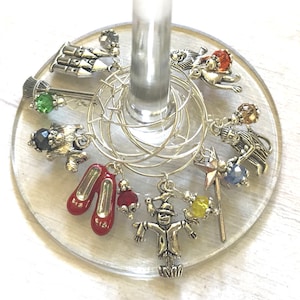 May include: A collection of silver-toned wine charms featuring iconic characters and symbols from The Wizard of Oz. Charms include ruby slippers, a scarecrow, a lion, and a wizard's wand. The charms are attached to a silver ring, designed to hang on a wine glass.