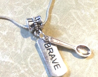 May include: Silver-toned necklace with a pendant featuring the word "BRAVE" on a rectangular charm and a miniature spoon charm. The necklace has a delicate chain and a decorative bead.