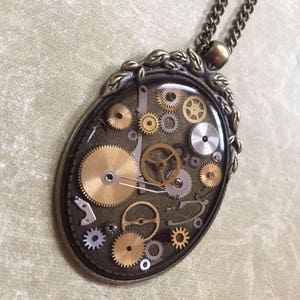 May include: Oval pendant necklace featuring a collection of watch gears in shades of gold and silver, encased in clear resin. The pendant has a bronze-colored frame with a decorative leaf design and hangs from a matching chain.