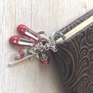 May include: A silver charm with red ballet slippers, a wand, and a red bead, attached to a brown leather-bound book. The charm is detailed and decorative, perfect for a book lover.
