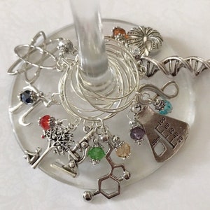 May include: A collection of silver wine glass charms with science-themed pendants. Charms include an atom, DNA strand, Erlenmeyer flask, and molecular structure, each adorned with colorful beads. The charms are attached to silver rings.
