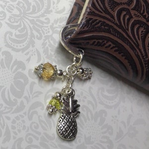 May include: Silver-toned bookmark charm with a cross design, featuring a yellow bead and a small pineapple charm. The charm is attached to a brown leather-bound book with an embossed floral pattern. The background is a light gray floral pattern.