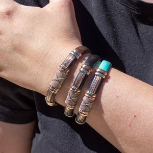 Puede incluir: Tres pulseras de cuero con detalles en cobre y plata. Las pulseras est&aacute;n apiladas en la mu&ntilde;eca de una persona.