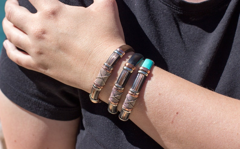 Puede incluir: Tres pulseras de cuero con detalles en cobre y plata. Las pulseras est&aacute;n apiladas en la mu&ntilde;eca de una persona.