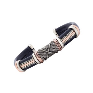 Puede incluir: Un brazalete de cuero negro con detalles plateados y cobrizos. El brazalete tiene un dise&ntilde;o tejido y est&aacute; acentuado con tres bandas de cobre.