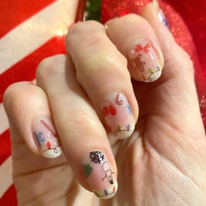 May include: Close-up of a hand with Christmas-themed nail art. Each nail features a different design, including a snowman, a gingerbread man, candy canes, and snowflakes. The designs are surrounded by a string of white lights.