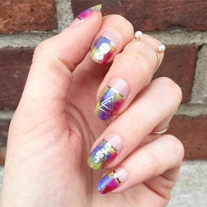 May include: Close-up of a hand with colorful nail polish with silver geometric designs. The nails are oval shaped and the polish is a mix of purple, blue, yellow, and green.