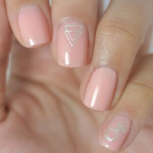 May include: Close-up of a hand with nude pink nail polish and gold geometric nail art designs. One nail has a triangle design, and another nail has a ring design.