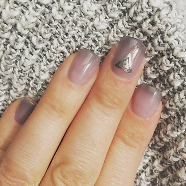 May include: Close-up of a hand with ombre nail polish. The ring finger nail has a silver triangle design.