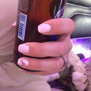 May include: A hand with pink nail polish and a ring on the ring finger holds a brown glass bottle. The bottle has a barcode and the text "love" is written on the finger.